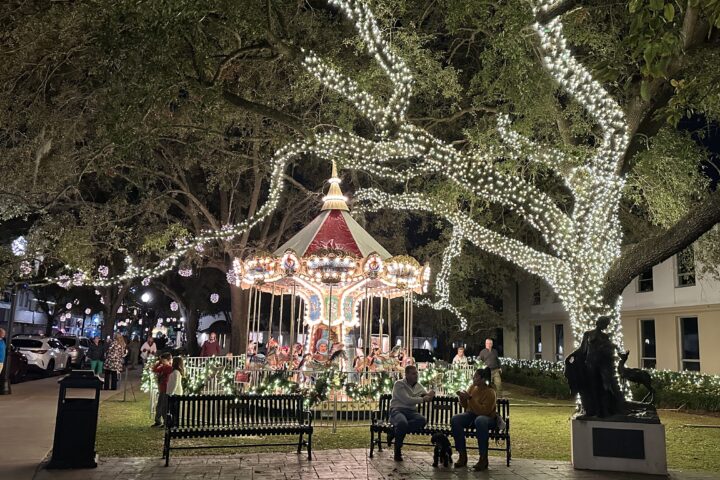 lighted carousel under large oak tree with park benches in front and people smiling all around
