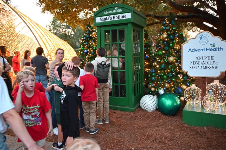 Colorful holiday scene at Winter Park's Christmas event featuring a green Santa hotline booth, decorated Christmas trees, and children waiting in line, showcasing festive community celebrations.