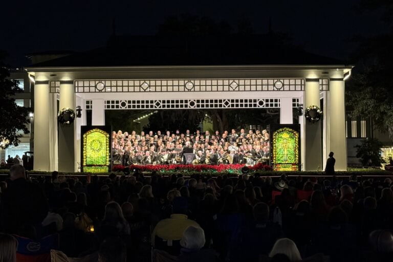 Outdoor concert at the Winter Park Community Band Shell during the evening, featuring a large choir and orchestra performing for a gathered audience in a festive, well-lit park setting.
