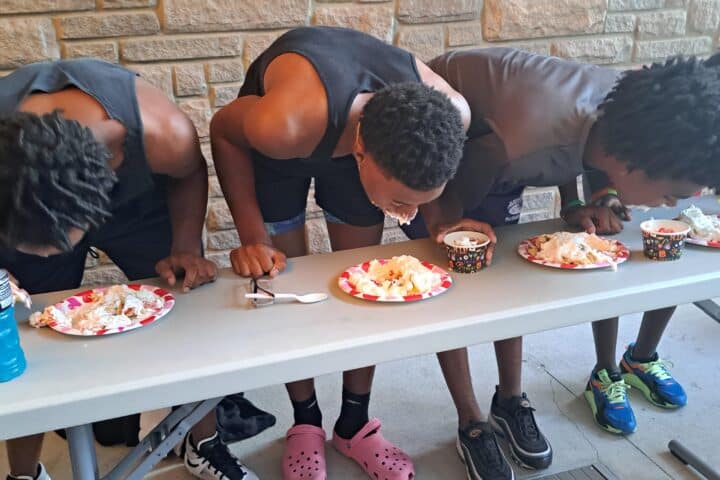 three boys standing behind a table bending down eating pie without their hands.