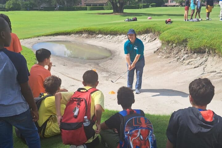 A group of children sitting on the grass and sand trap at Winter Park golf course, learning golf skills from an instructor outdoors on a sunny day, in the scenic city park environment.