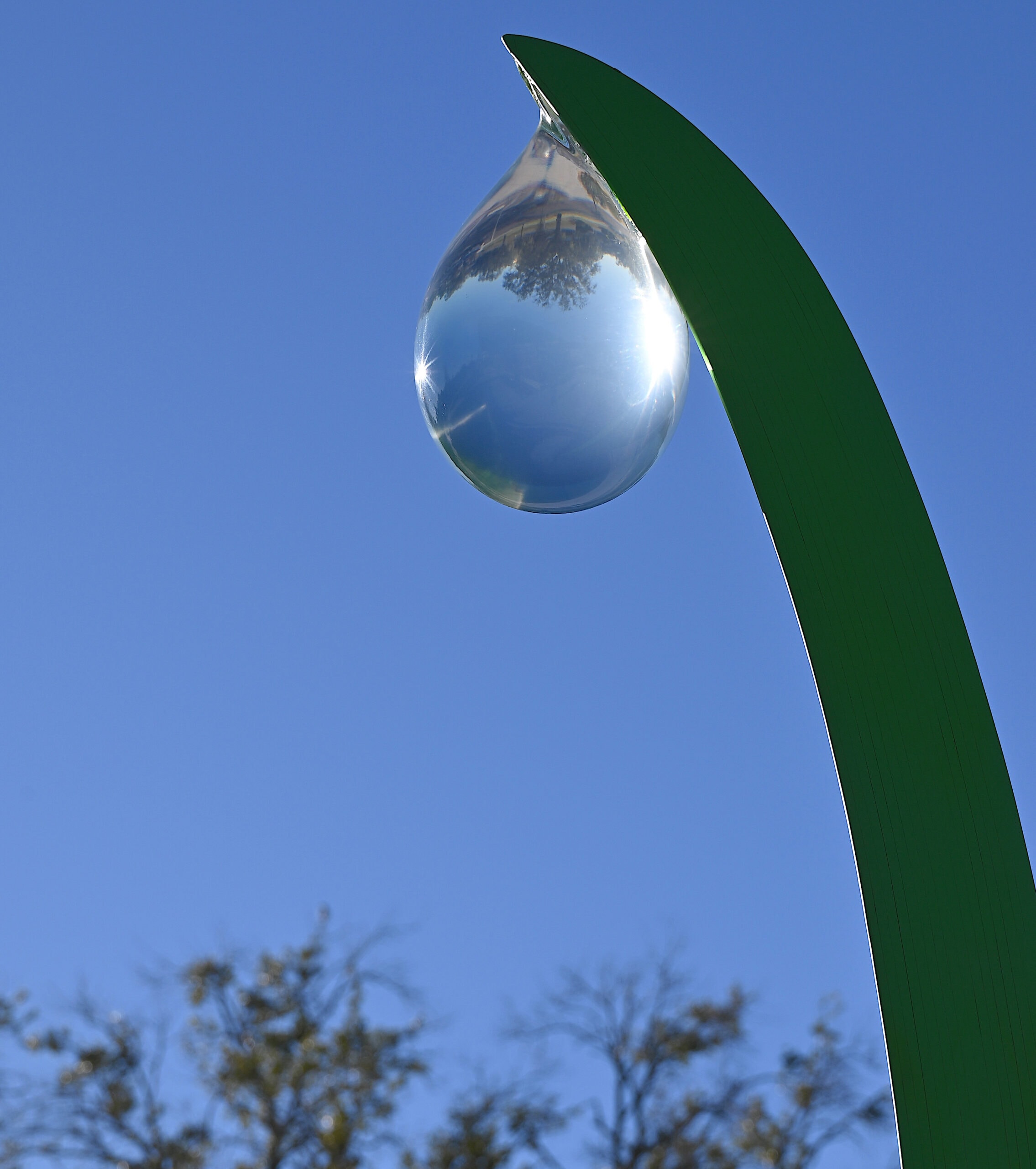 sculpture of water droplet with reflection of outdoor surroundings hanging from metal blade of grass