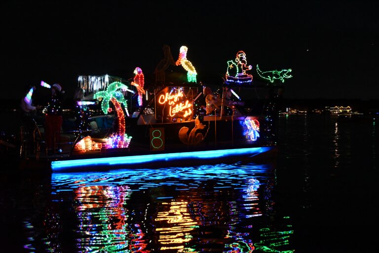 A boat cruising at night, brightly decorated with colorful holiday lights. Lighted figures include palm trees, pelican, parrot, gator and a glowing sign.
