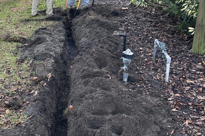 Workers dig a narrow trench beside a lawn for utility installation.