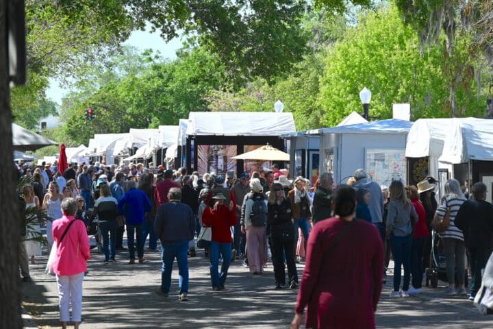A large crowd of people walk along a tree-lined art festival at the Winter Park Sidewalk Art Festival, with white tented booths on both sides displaying artwork. The lively, sunny atmosphere highlights this vibrant event held March 20-22.