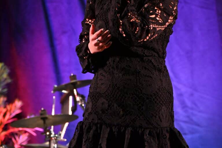 A woman in a black lace dress stands on stage holding a microphone, performing in front of colorful, softly lit curtains and a drum set in the background.
