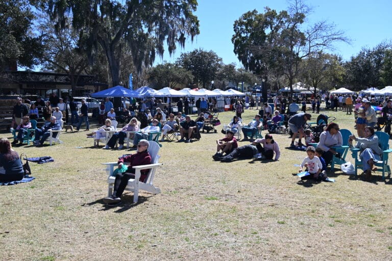 People sit on lawn chairs and picnic blankets in a grassy park area during a sunny outdoor event, with many vendor tents and trees in the background. Some attendees are eating, chatting, or relaxing.