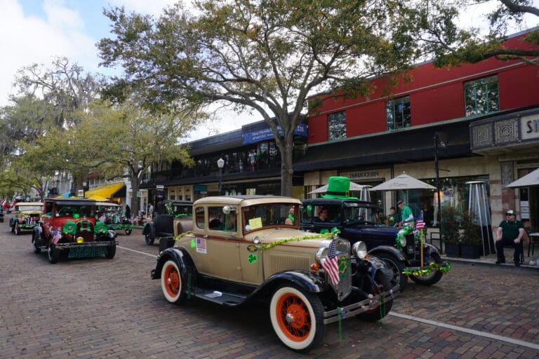 A parade of vintage cars decorated with green shamrocks and flags drives down a brick street lined with trees and shops, celebrating St. Patrick’s Day. People watch from the sidewalks and sit at outdoor tables.