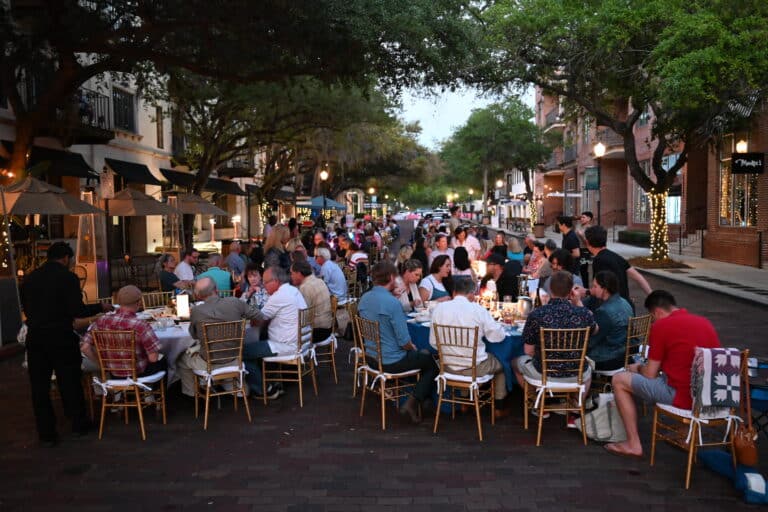 People dining at long tables set up on a closed street lined with trees and twinkling lights in the evening.