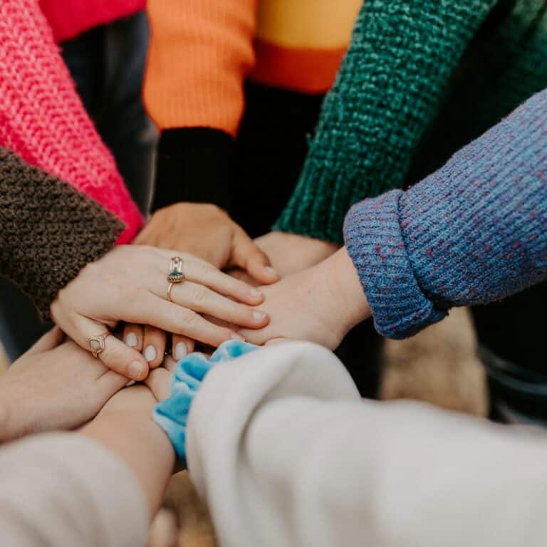 A group of people wearing colorful sweaters stack their hands, celebrating unity and the simple things to enjoy together.