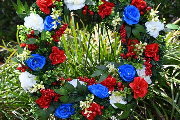 A circular wreath with red, white, and blue flowers on a metal stand, displayed outdoors in a garden.