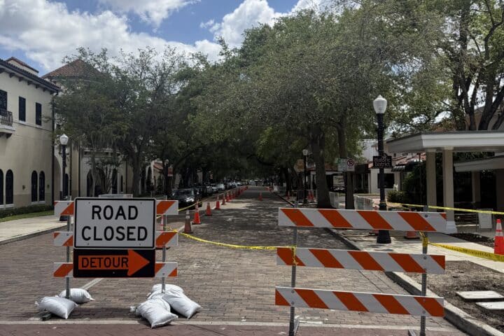 A brick street blocked by Road Closed signs, orange cones, and caution tape under a sunny sky.