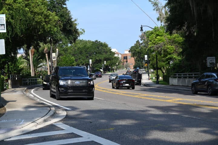 Several cars drive on SR426, a curving road lined with trees and FDOT signs, on a sunny day.