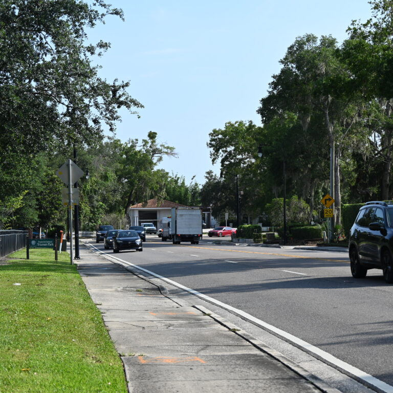 Cars driving on a suburban street lined with grass, trees, and sidewalk on a sunny day.