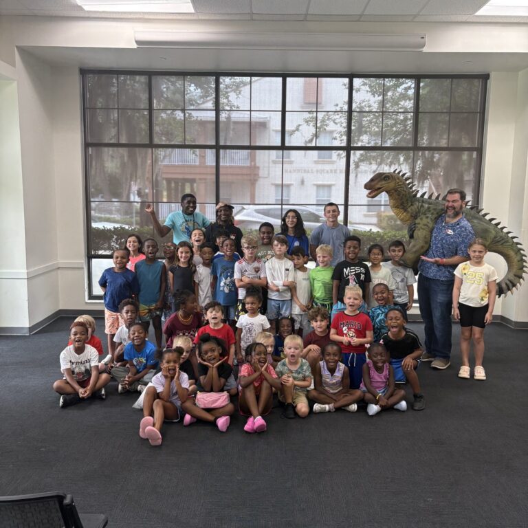A group of children and adults pose indoors during student holidays with a person in a dinosaur costume in the background.