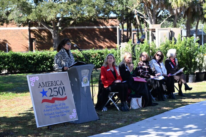 A speaker addresses attendees at an outdoor America 250! event hosted by the Daughters of the American Revolution.