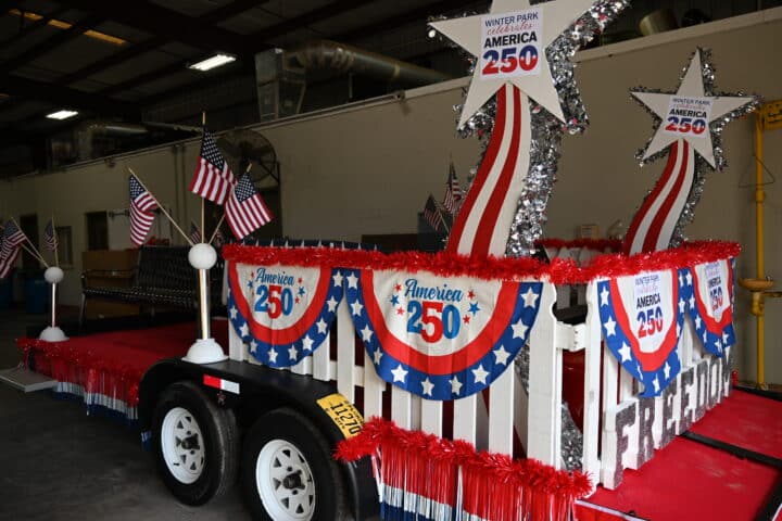 Patriotic parade float decorated with American flags, stars, and America 250 banners inside a warehouse.