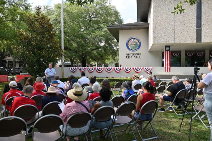 People sit outside Winter Park City Hall for an event, with patriotic bunting and flags decorating the building.