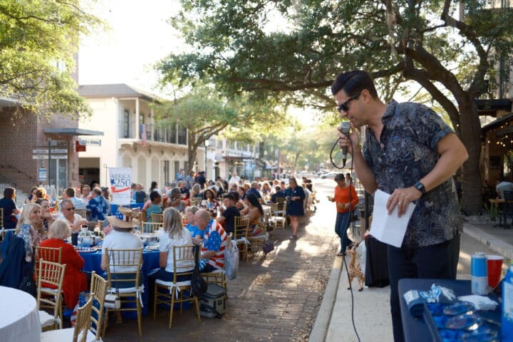 A man speaks into a microphone at an outdoor event with many people seated at tables under trees.