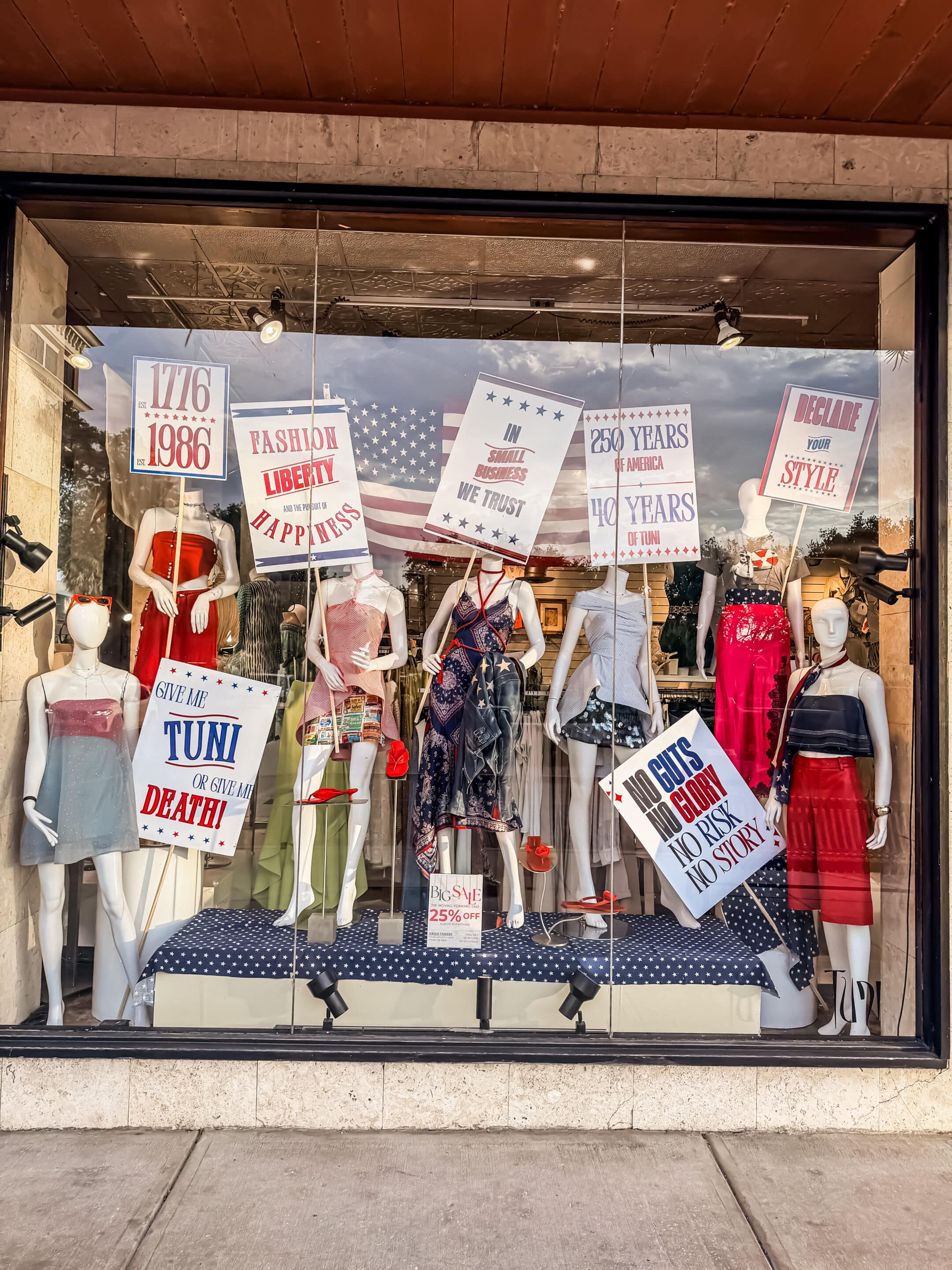 Storefront display with mannequins in patriotic outfits, decorated with red, white, and blue Independence Day signs.