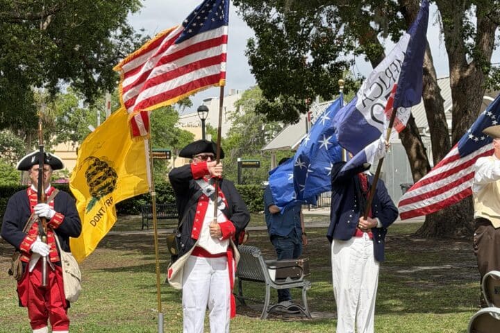 People in historical military uniforms hold flags, including the U.S. flag and Dont Tread on Me, in a park.