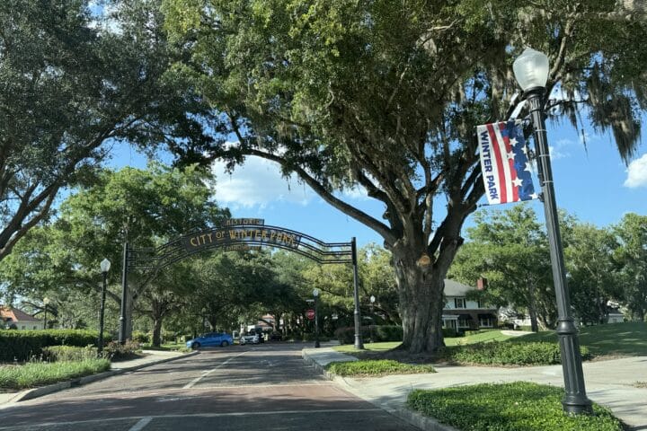 A tree-lined street with a City of Winter Park archway and a Winter Park patriotic banner on a lamp post.
