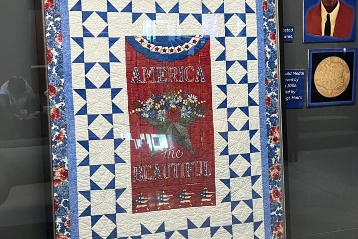 A framed quilt with a red center reading America the Beautiful, bordered by blue and white patterns and red flowers.
