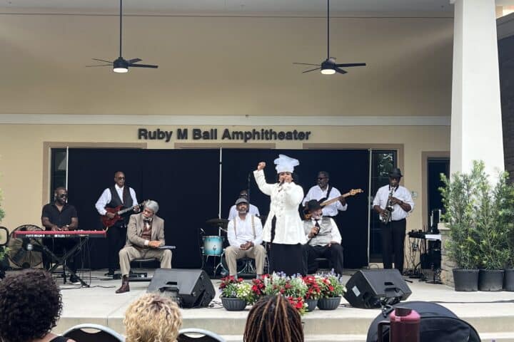 A band performs on stage at Ruby M Ball Amphitheater, with a singer in white and audience members watching.