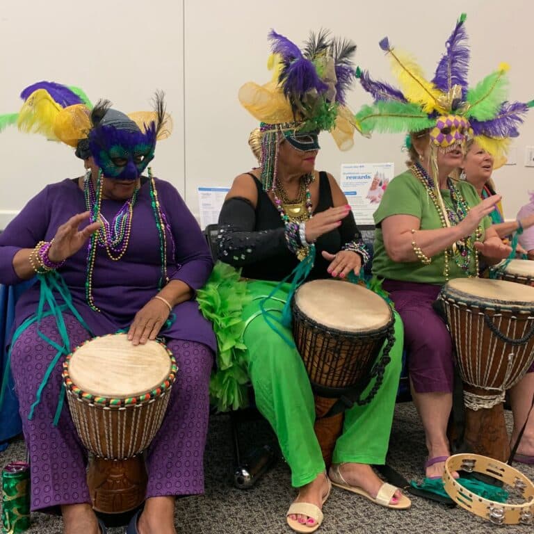 People in colorful masks and costumes play drums together at a vibrant Community Center celebration indoors.