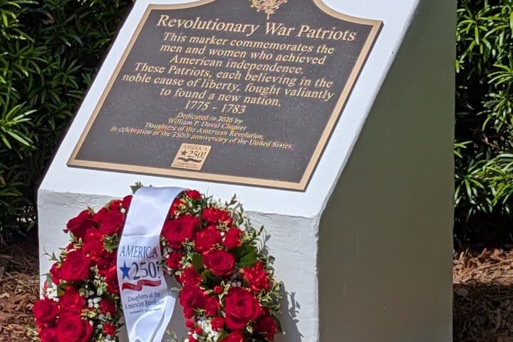 A memorial plaque for Revolutionary War Patriots with a wreath of red roses at its base, set outdoors among greenery.