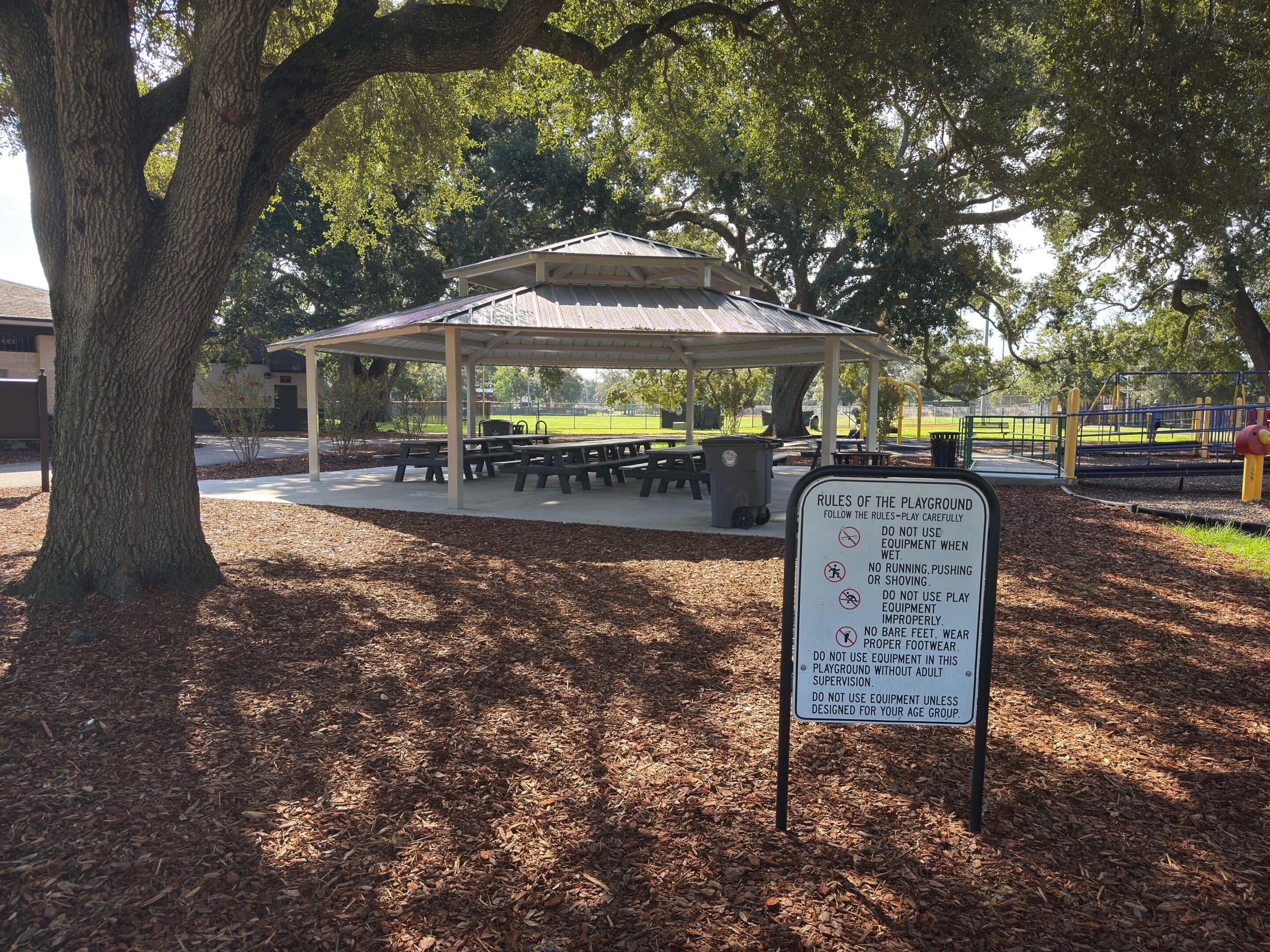 A shaded picnic pavilion with tables stands near a playground; a sign lists playground rules in the foreground.